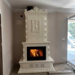 Usługi Zduńskie Piotr Oskwarek - Kremowy kominek kaflowy z paleniskiem, dekoracyjnymi zdobieniami i wzorem na podstawie. Widoczne płomienie w palenisku. Na podłodze rozłożony papier zabezpieczający.