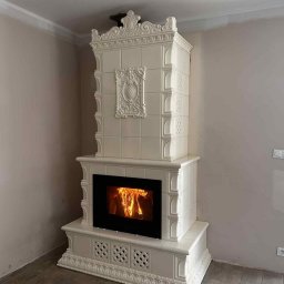 Usługi Zduńskie Piotr Oskwarek - Biały, kaflowy kominek z paleniskiem, zdobiony ornamentami, w jasnym wnętrzu. Widoczne płomienie i dekoracyjne wykończenie. Styl tradycyjny, kafle ułożone w kwadraty.