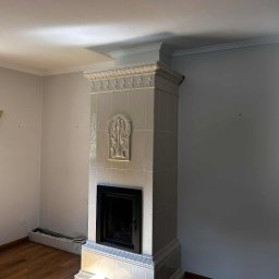 Usługi Zduńskie Piotr Oskwarek - Biały, kaflowy kominek z dekoracyjnymi elementami w jasnym wnętrzu. Widoczne drewniane podłogi i fragment ściany. Kominek w stylu retro z ozdobnymi kaflami.