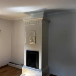 Biały, kaflowy kominek z dekoracyjnymi elementami w jasnym wnętrzu. Widoczne drewniane podłogi i fragment ściany. Kominek w stylu retro z ozdobnymi kaflami.