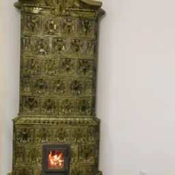 Zdobiony, oliwkowy piec kaflowy w rogu pokoju, z palącym się ogniem w palenisku. Widoczne detale ornamentów na kaflach.