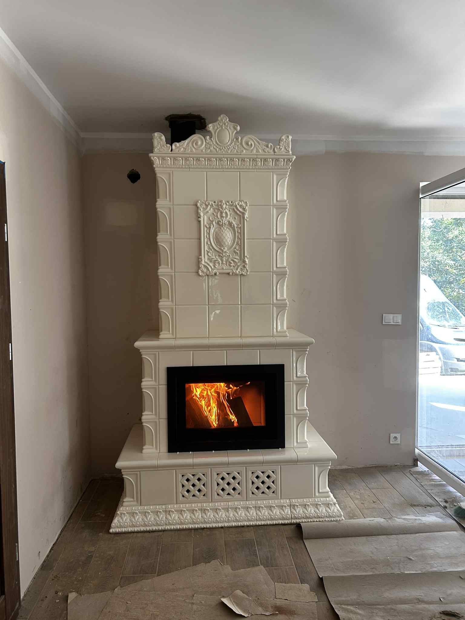 Kremowy kominek kaflowy z paleniskiem, dekoracyjnymi zdobieniami i wzorem na podstawie. Widoczne płomienie w palenisku. Na podłodze rozłożony papier zabezpieczający.