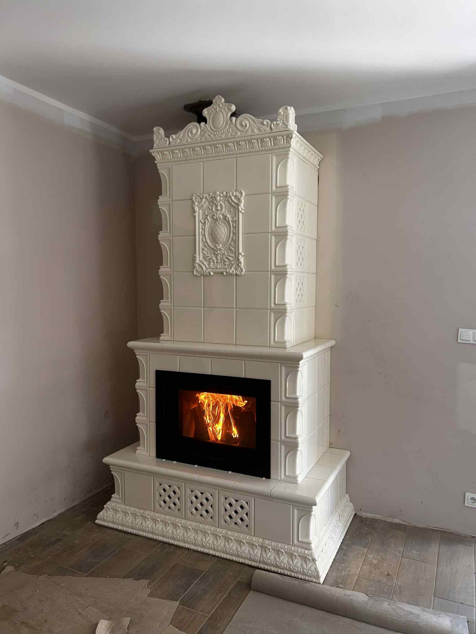 Biały, kaflowy kominek z paleniskiem, zdobiony ornamentami, w jasnym wnętrzu. Widoczne płomienie i dekoracyjne wykończenie. Styl tradycyjny, kafle ułożone w kwadraty.