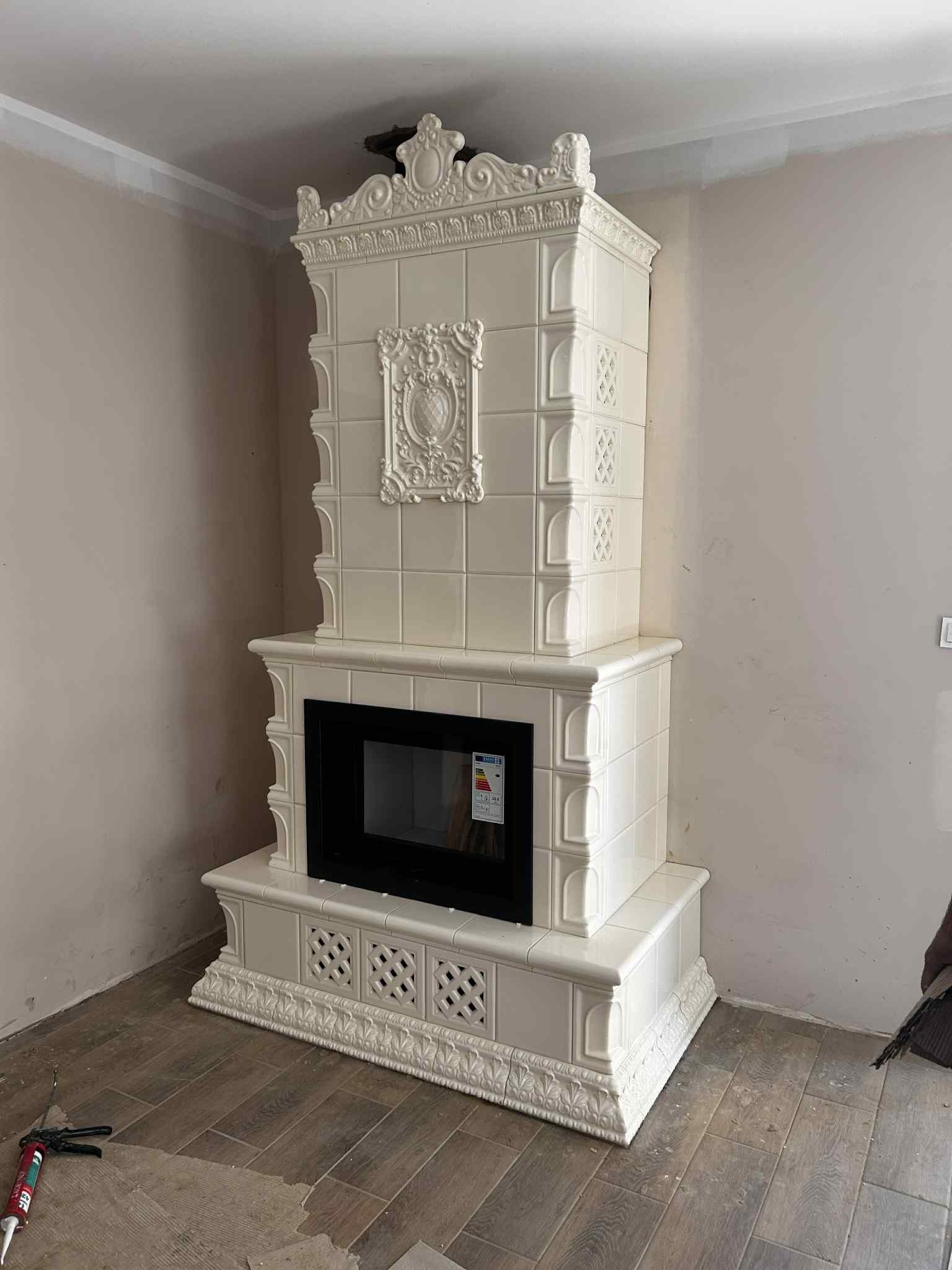 Biały, kaflowy kominek z czarnym wkładem kominkowym, zdobiony ornamentami, w trakcie montażu w Kolsku. Na podłodze widoczne narzędzia i płytki.