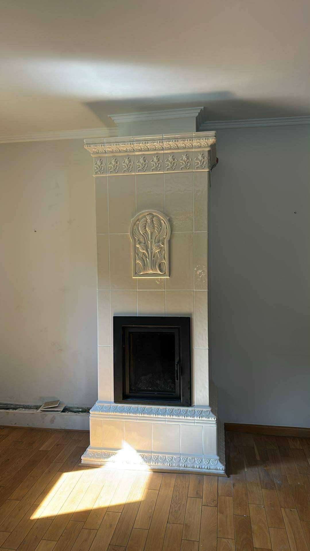 Biały piec kaflowy z czarnymi drzwiczkami, zdobiony ornamentami roślinnymi, w jasnym wnętrzu z parkietem. Widoczny detal wykończenia i dekoracyjny charakter pieca.