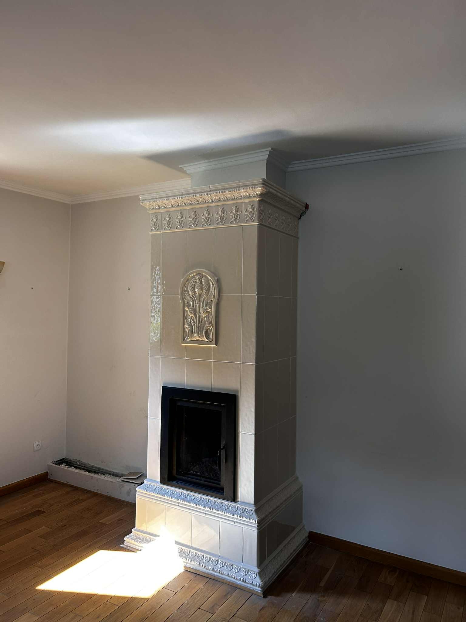 Biały, kaflowy kominek z dekoracyjnymi elementami w jasnym wnętrzu. Widoczne drewniane podłogi i fragment ściany. Kominek w stylu retro z ozdobnymi kaflami.