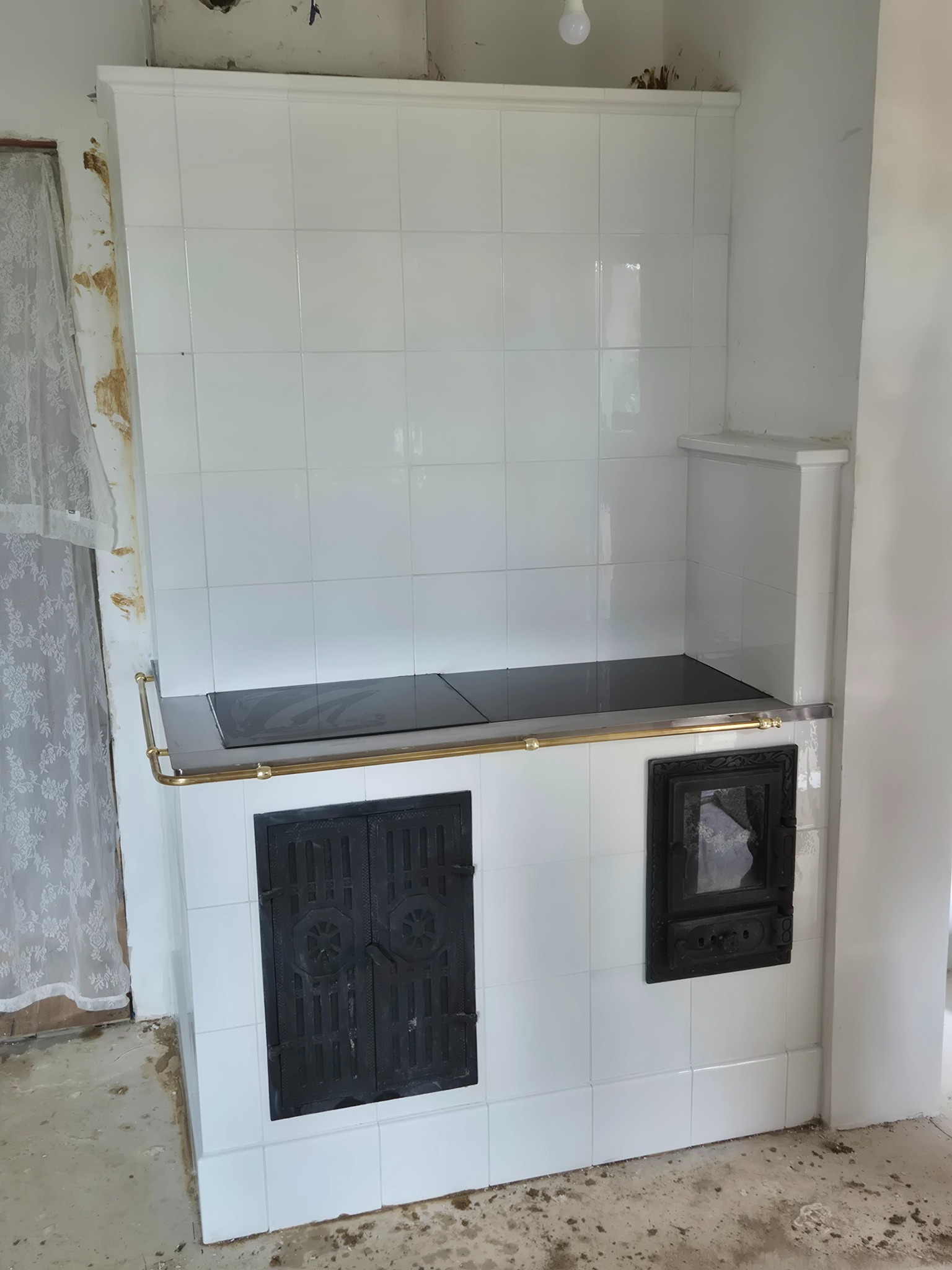 Biała, kafelkowa kuchnia kaflowa z czarną płytą grzewczą i drzwiczkami paleniska. Widoczne niedokończone wykończenie ściany i podłogi. Oświetlenie górne.