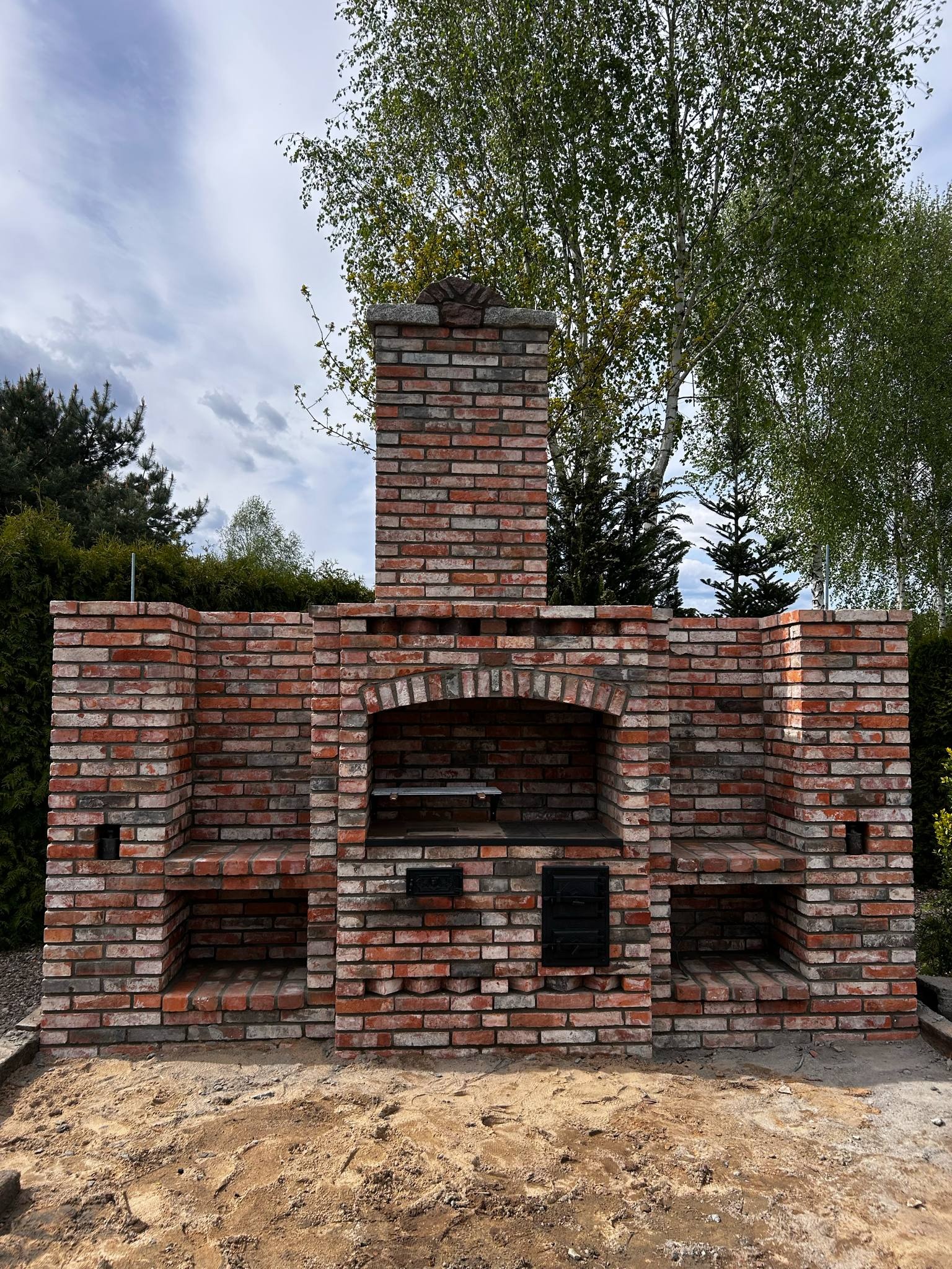 Murowany grill ogrodowy z cegły rozbiórkowej, z płytą kuchenną i słupkami podtrzymującymi konstrukcję dachową. Otoczenie zieleni, pochmurne niebo.