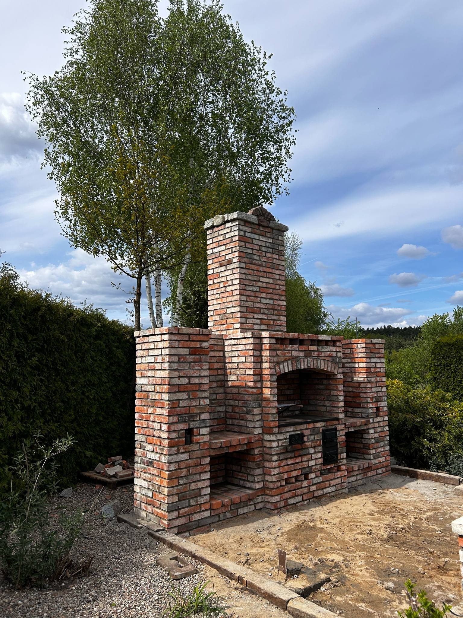 Murowany grill ogrodowy z cegły klinkierowej na tle zieleni i błękitnego nieba z chmurami. Widoczne elementy konstrukcyjne i otoczenie.