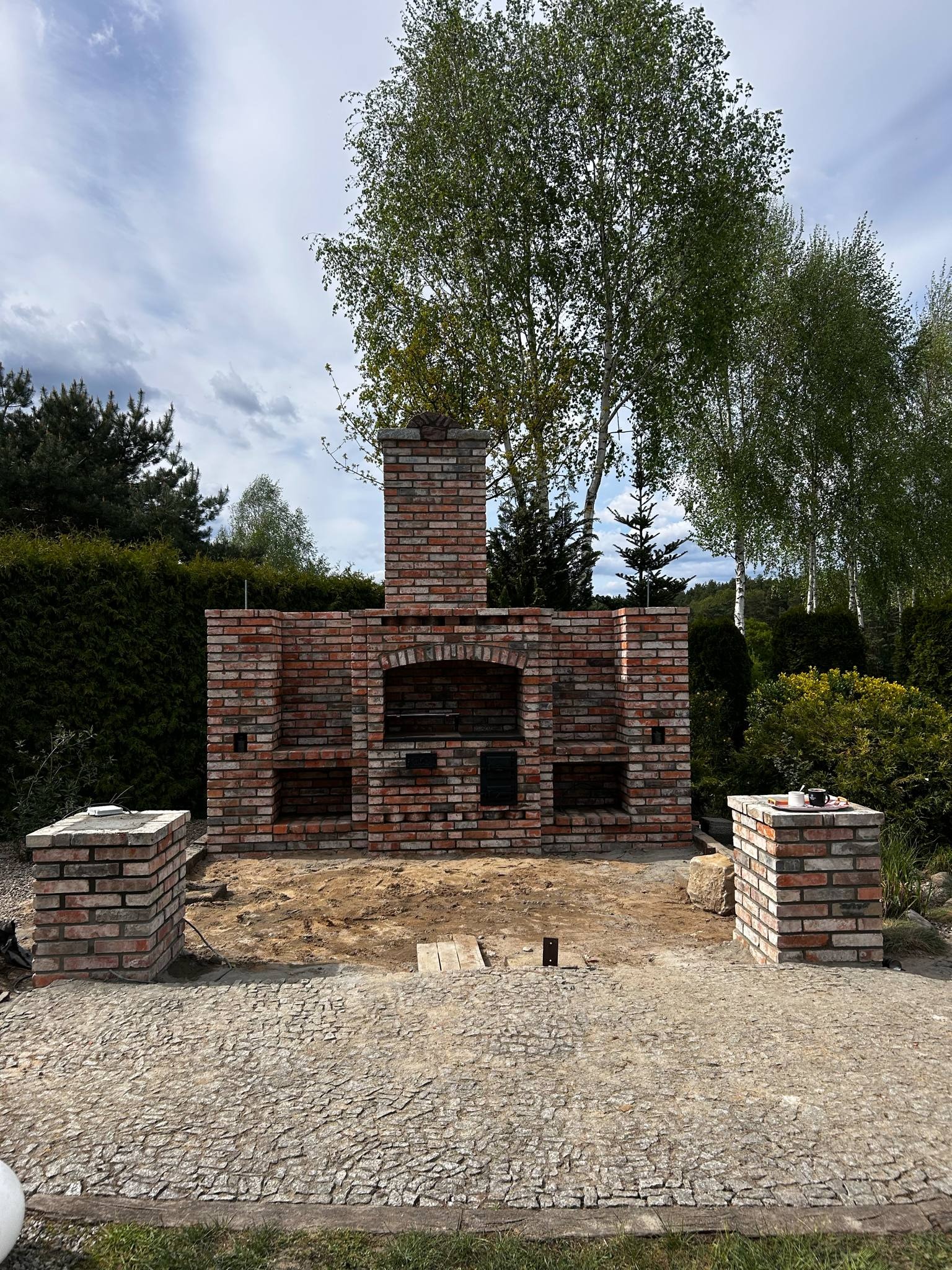 Murowany, ceglany grill ogrodowy w trakcie budowy, z ceglanym kominem i dwoma filarami. Na jednym z filarów kawa i ciastko. W tle drzewa i niebo.