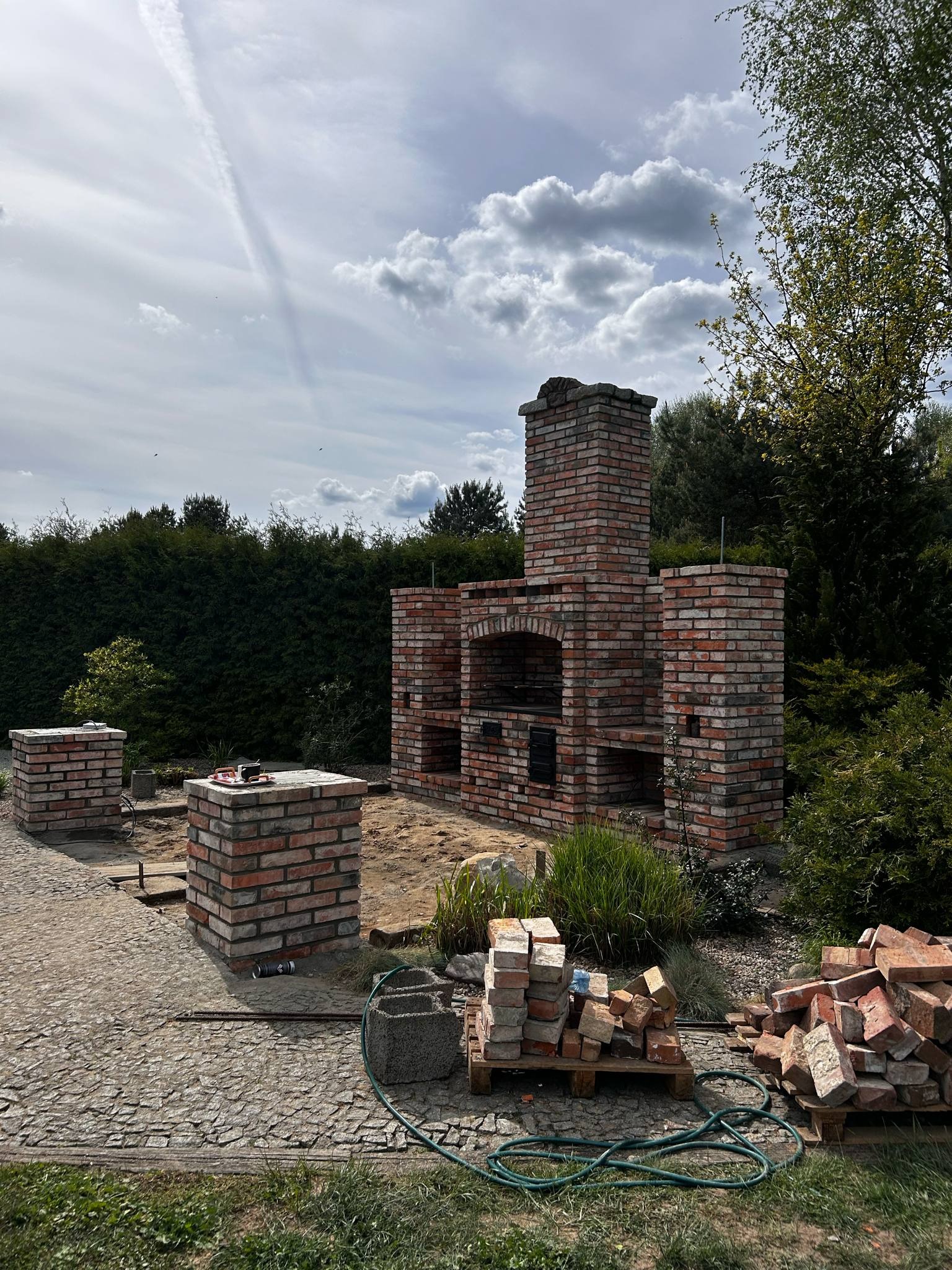 Murowany grill ogrodowy z cegły w trakcie budowy. Widoczne materiały budowlane i elementy konstrukcyjne. Tło: zieleń, pochmurne niebo.