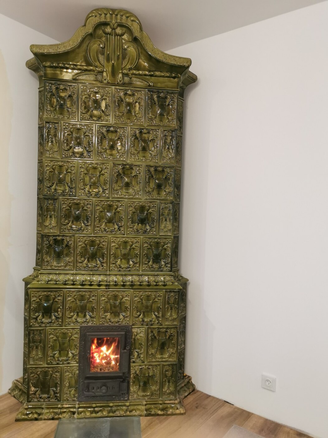 Zdobiony, oliwkowy piec kaflowy w rogu pokoju, z palącym się ogniem w palenisku. Widoczne detale ornamentów na kaflach.