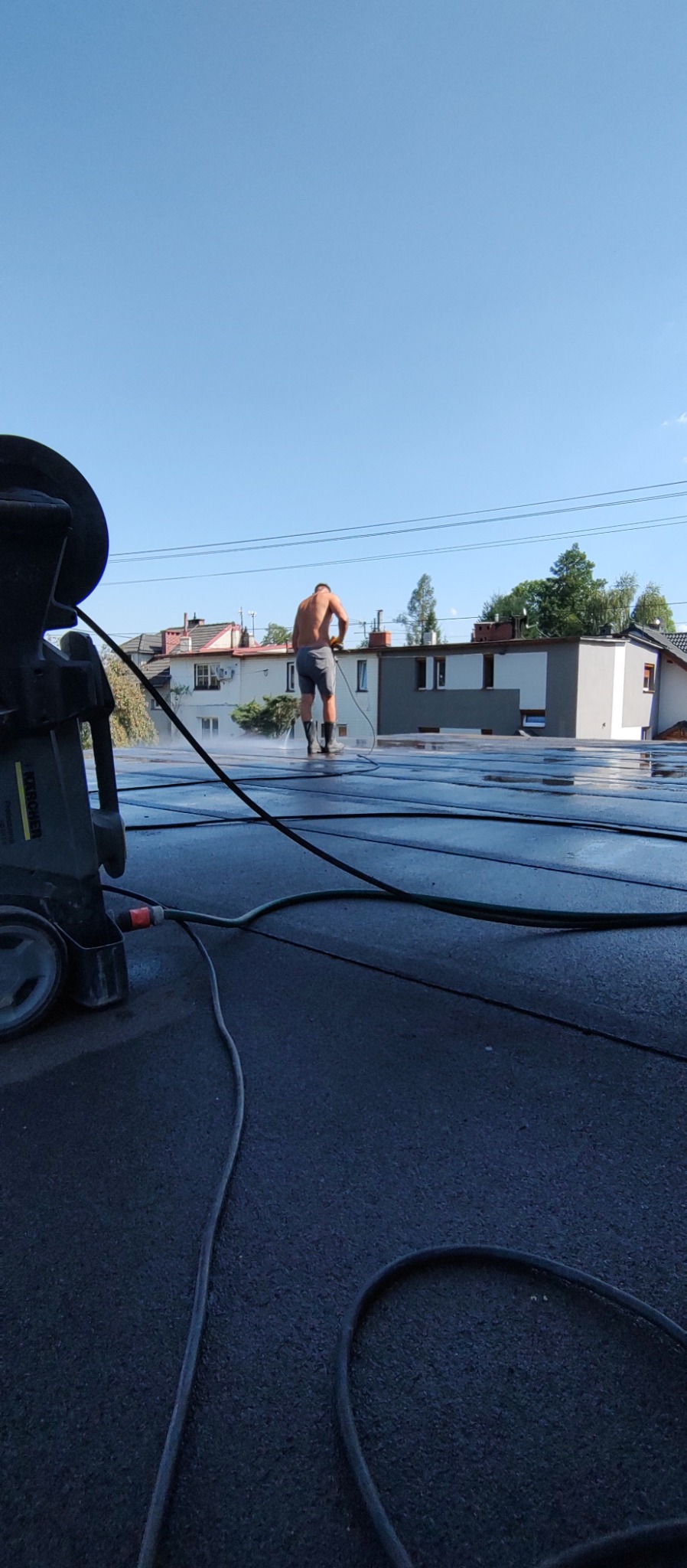 Mężczyzna w krótkich spodenkach i kaloszach czyści płaski dach myjką ciśnieniową, widoczne przewody i urządzenie czyszczące.