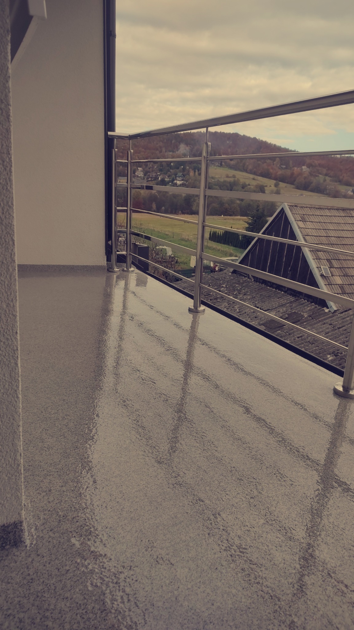 Balkon z szarą, połyskującą posadzką żywiczną typu dywan kwarcowy i metalową balustradą, widok na pagórkowaty krajobraz z zabudowaniami.