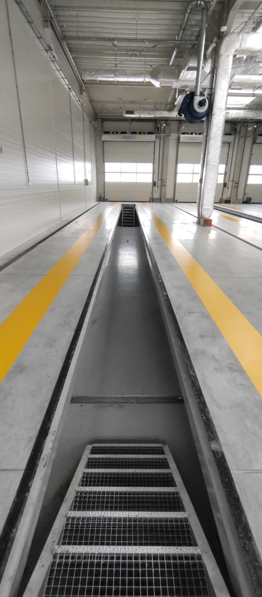 Wnętrze hali przemysłowej z kanałem rewizyjnym i żółtymi pasami na posadzce. Widoczne schody zejściowe do kanału i brama wjazdowa w tle.