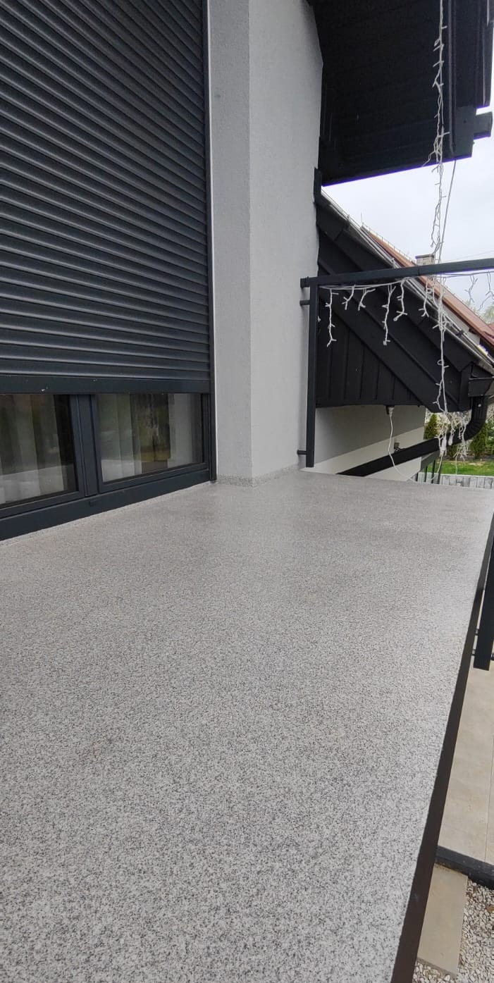 Jasnoszara, drobnoziarnista posadzka balkonu z widokiem na okno z roletą i fragment domu z dekoracyjnymi lampkami. Nowoczesne wykończenie.