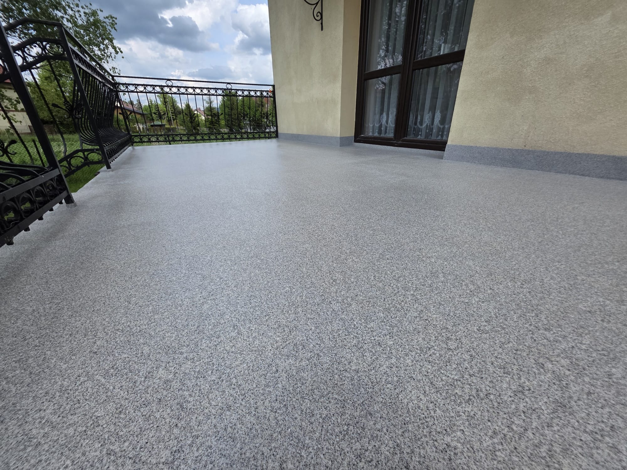 Balkon z szarą, wzorzystą posadzką żywiczną, czarną, ozdobną balustradą i beżową elewacją. Widok na zieleń i niebo z chmurami. Nowoczesne wykończenie.