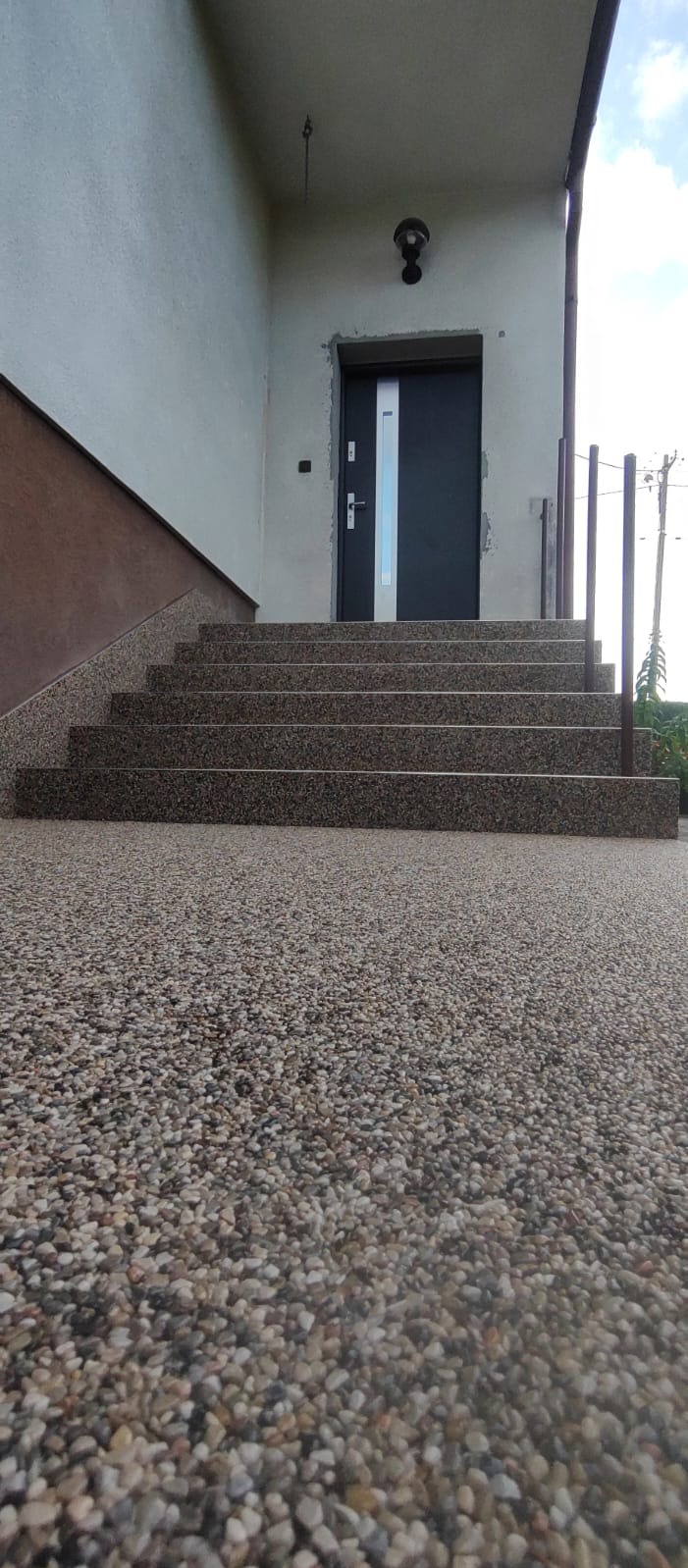 Schody wejściowe wykończone drobnym kamieniem w żywicy, prowadzące do ciemnych drzwi z metalową listwą. Widoczne barierki i fragment elewacji budynku.