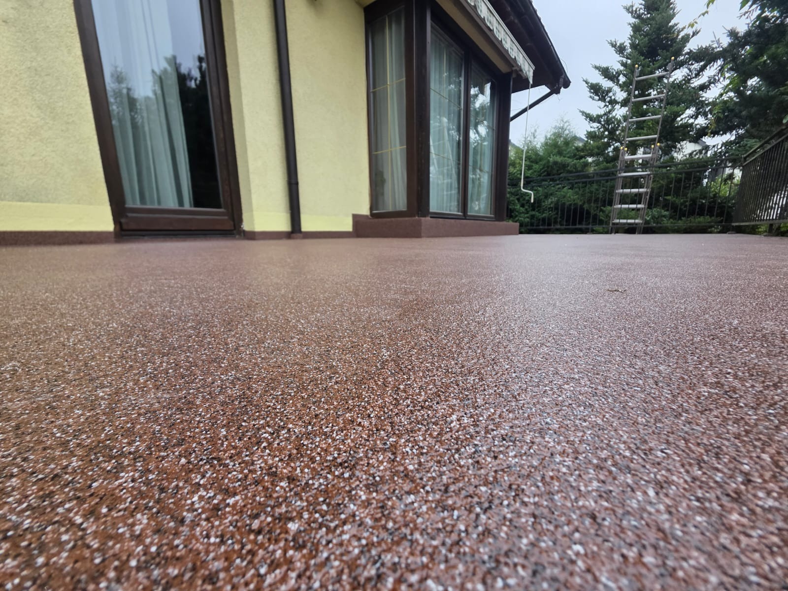 Balkon z brązową posadzką żywiczną z białymi drobinkami, widok z dołu. W tle ściana budynku z oknami i drabina oparta o drzewo.