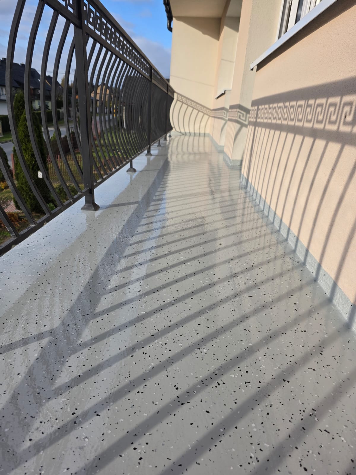 Szara, błyszcząca posadzka żywiczna na balkonie z czarną, metalową balustradą. Widoczne cienie balustrady i dekoracyjny wzór na elewacji budynku.