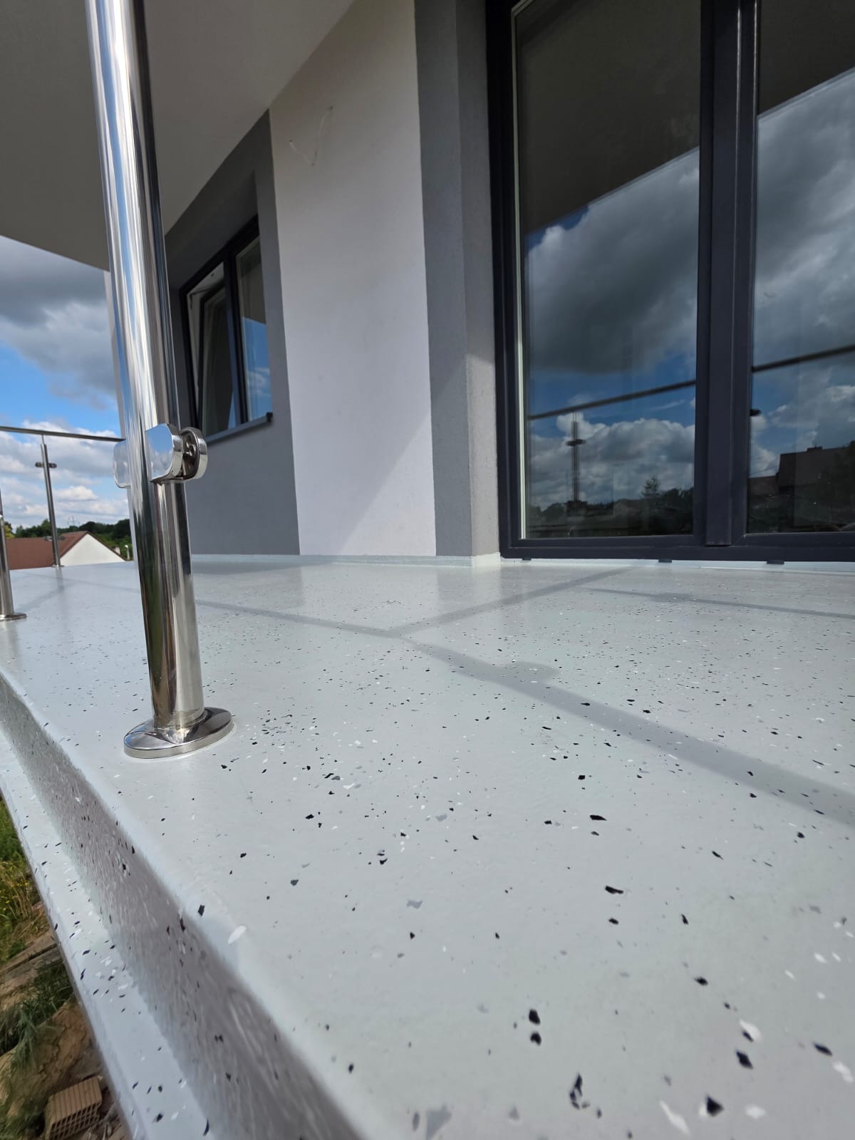 Szara posadzka żywiczna na balkonie z metalową balustradą i widokiem na okno z odbiciem chmur. Nowoczesne wykończenie balkonu.