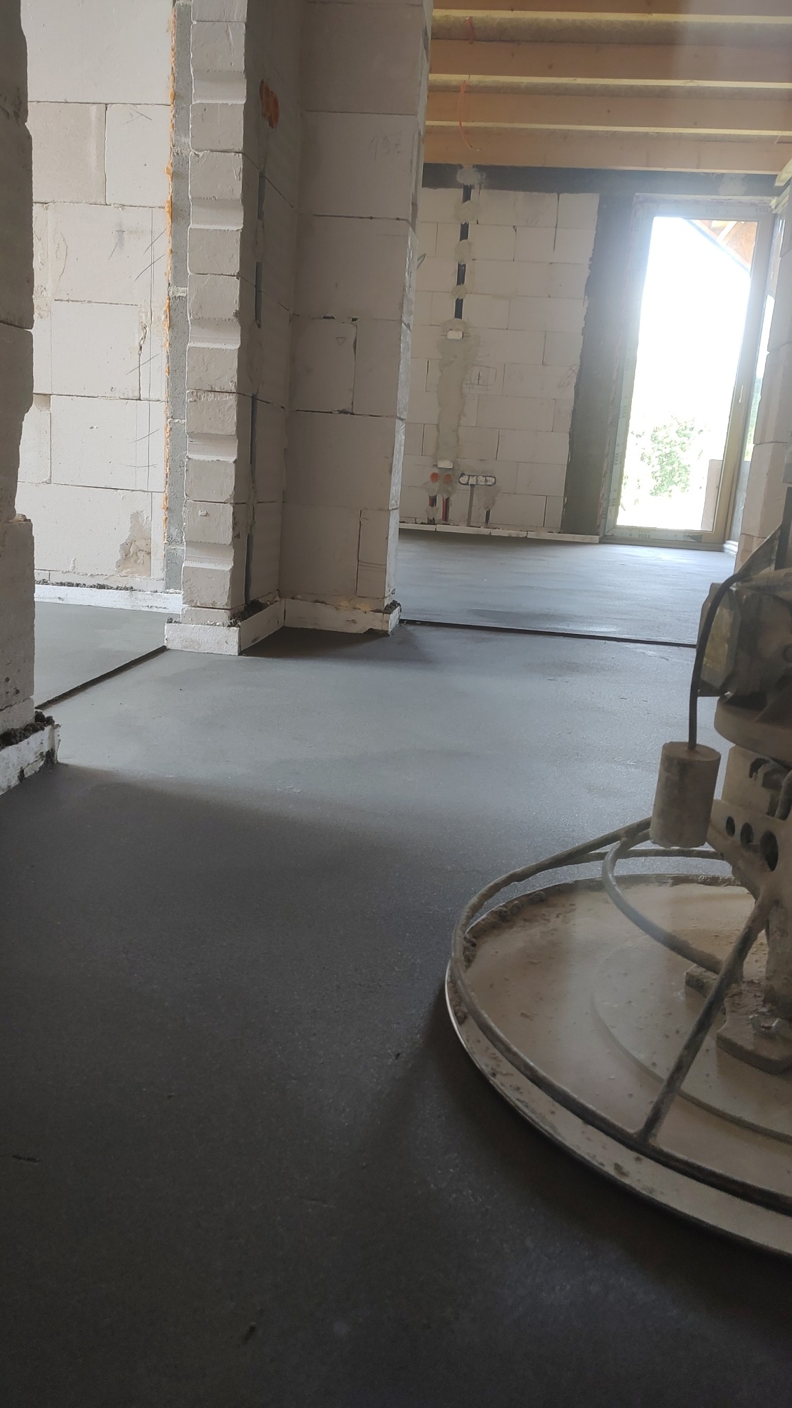 Wyrównana, szara posadzka jastrychowa w surowym wnętrzu budynku w trakcie budowy. Widoczna maszyna do zacierania. Świeżo wylana podłoga, jasne ściany z pustaków.