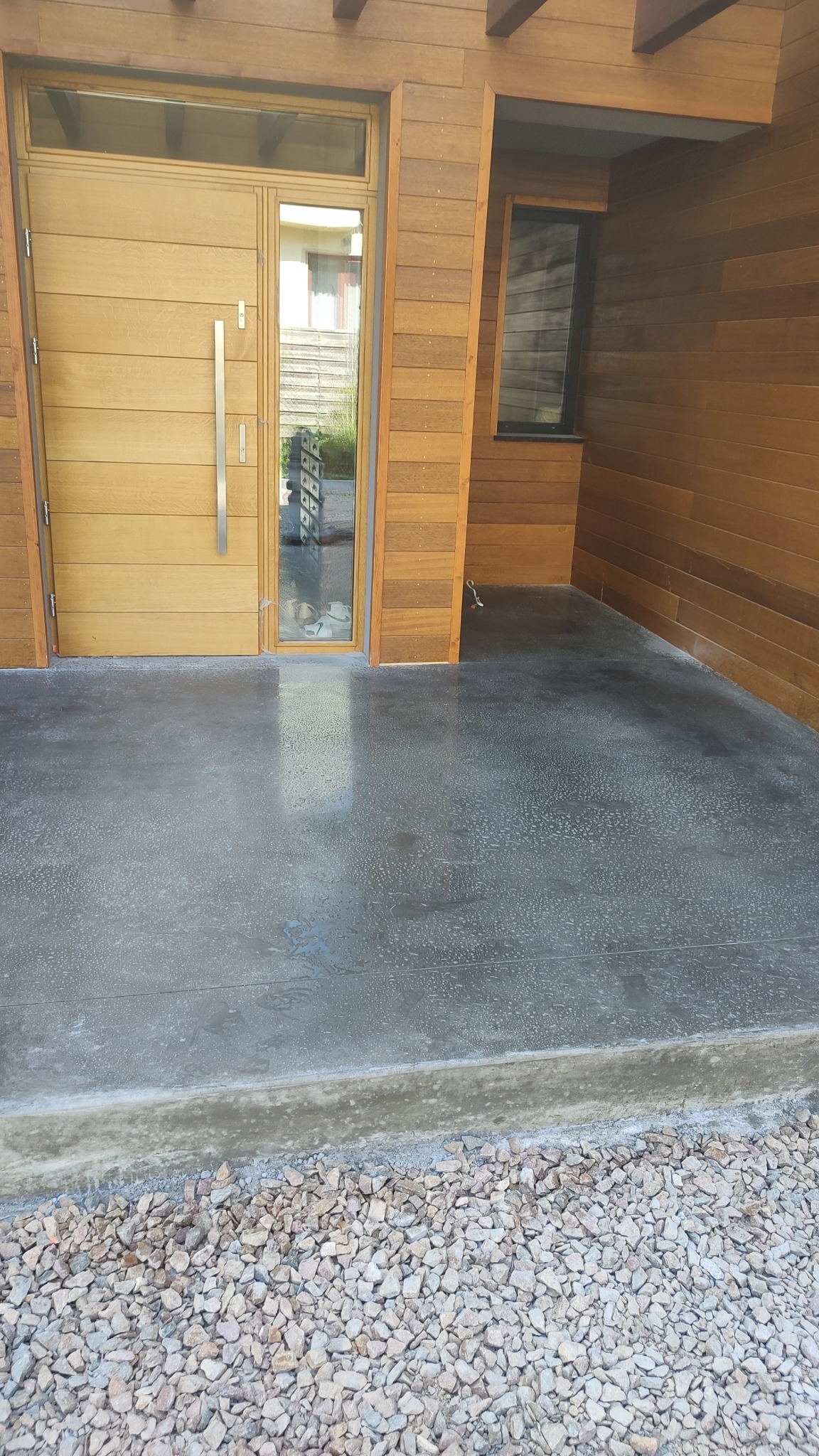 Szara, gładka posadzka na zewnątrz budynku z drewnianą elewacją i drzwiami. Widoczne kamienie ozdobne przy krawędzi posadzki. Minimalistyczny design.