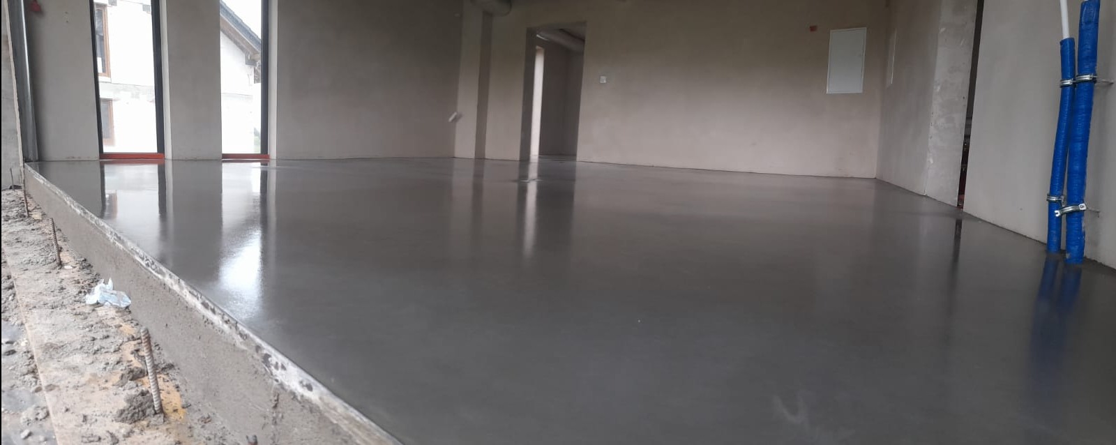 Świeżo wylana, gładka, szara posadzka w surowym wnętrzu z widocznymi instalacjami przy ścianie i oknami w tle. Widoczny fragment szalunku.