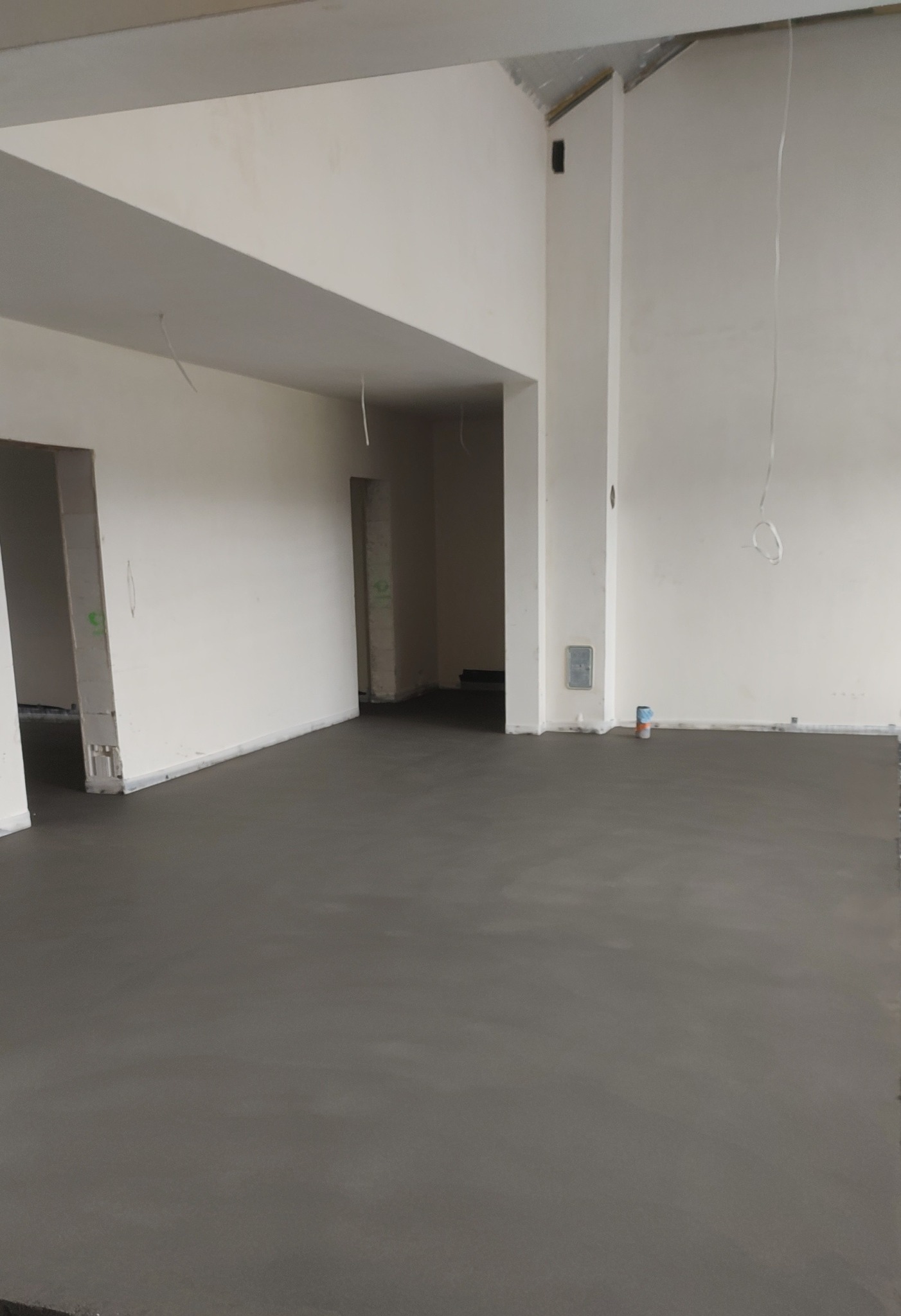Świeżo wylana, gładka posadzka jastrychowa w surowym wnętrzu z wysokim stropem. Widoczne kable zwisające z sufitu i puszka elektryczna na ścianie.