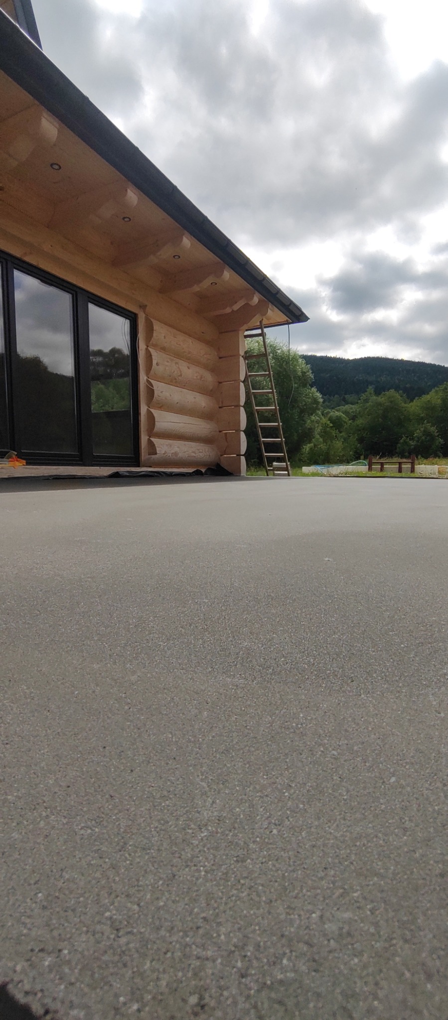 Szara, gładka posadzka balkonu drewnianego domu z widokiem na zalesione wzgórza w tle. Drabina oparta o ścianę. Ujęcie z niskiej perspektywy.