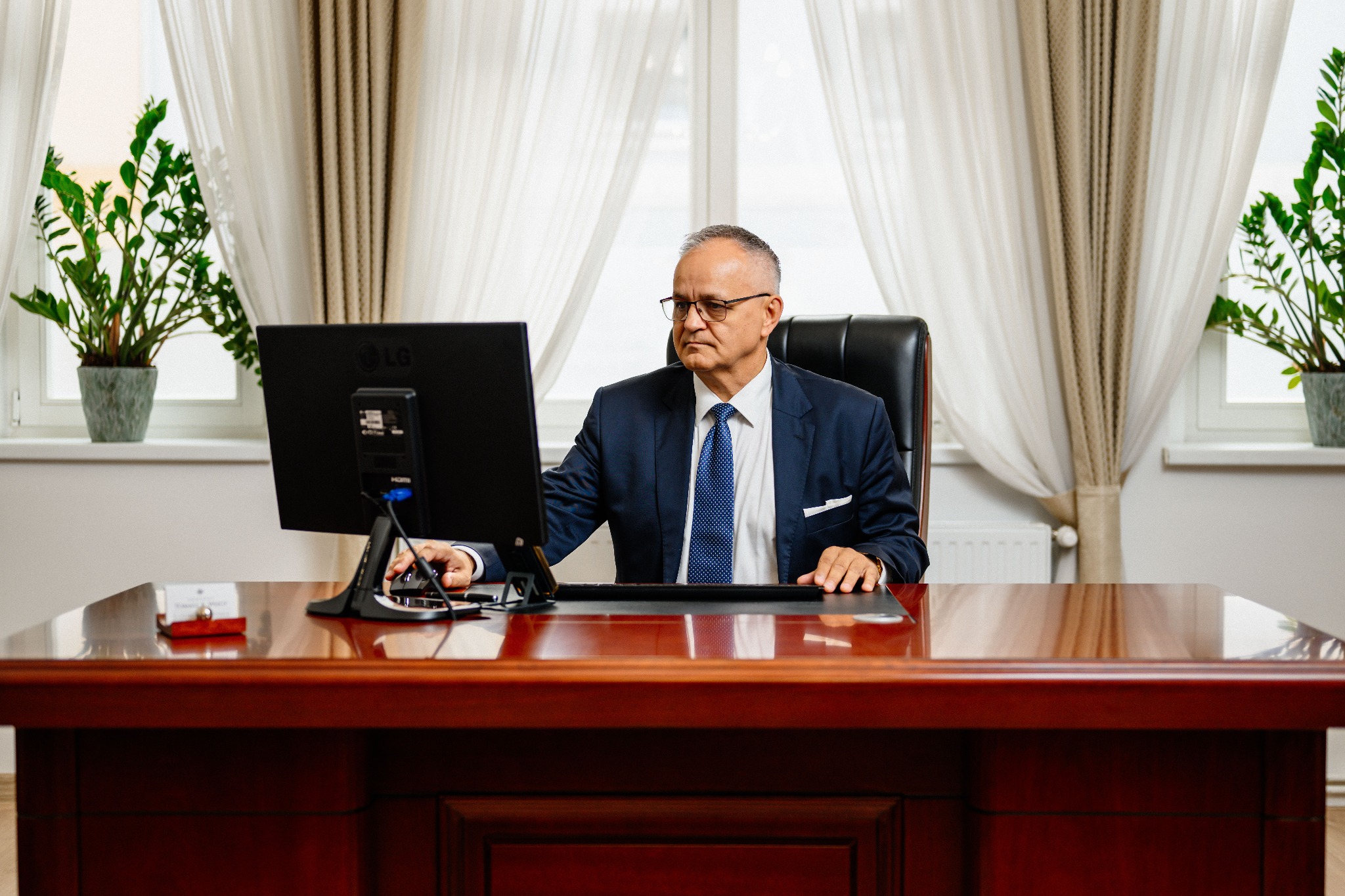 Adwokat w garniturze siedzi za biurkiem z komputerem, w tle okno z zasłonami i rośliny doniczkowe. Powaga i profesjonalizm w kancelarii.