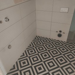 Alfa-Bud Andrzej Moczydlak - Łazienka w trakcie remontu: białe, poziome płytki ścienne z delikatnym wzorem, czarno-biała podłoga w geometryczne wzory, widoczne instalacje wodne i przycisk spłuczki.