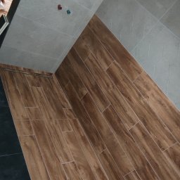 Alfa-Bud Andrzej Moczydlak - Wnętrze łazienki w trakcie prac wykończeniowych: ułożona podłoga z płytek imitujących drewno, szare płytki na ścianach, widoczne wystające rury instalacji wodnej oraz narzędzie do cięcia płytek.