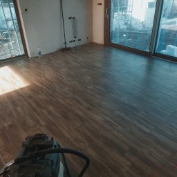 Alfa-Bud Andrzej Moczydlak - Układanie płytek imitujących drewno w jasnym pomieszczeniu z dużymi oknami, widoczna maszyna czyszcząca na podłodze i materiały budowlane za oknem.