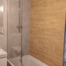Alfa-Bud Andrzej Moczydlak - Wykończona łazienka z wanną obłożoną płytkami imitującymi kamień, szklanym parawanem nawannowym oraz ścianą wyłożoną płytkami imitującymi drewno; w wannie leży słuchawka prysznicowa.