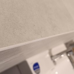 Alfa-Bud Andrzej Moczydlak - Szczegółowe ujęcie fugi między szarymi płytkami na ścianie w łazience, widoczny fragment umywalki z baterią i szafki pod umywalką.