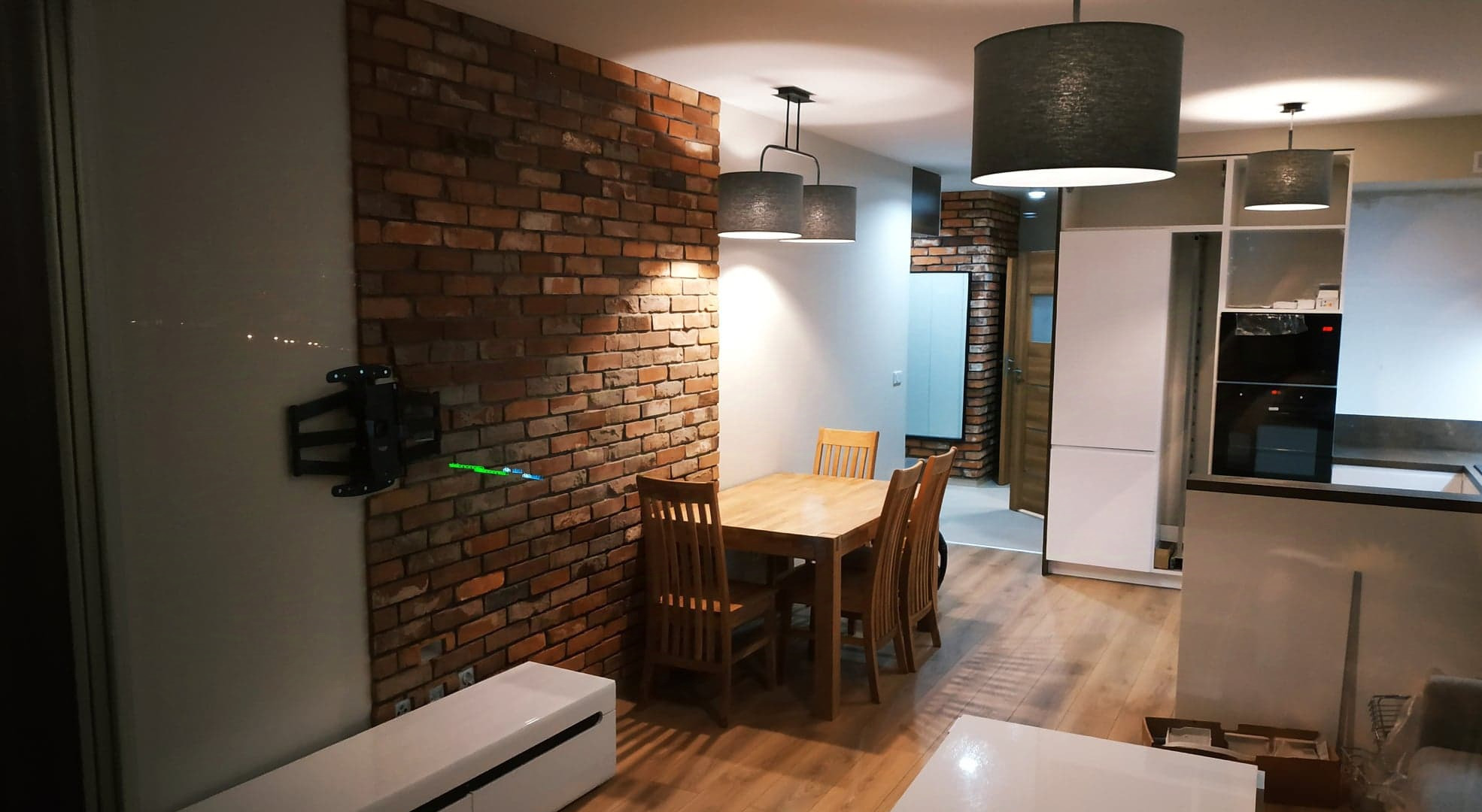 Jadalnia w stylu industrialnym z ceglaną ścianą, drewnianym stołem i krzesłami oraz wiszącymi lampami z abażurami, widoczny uchwyt do telewizora na ścianie.