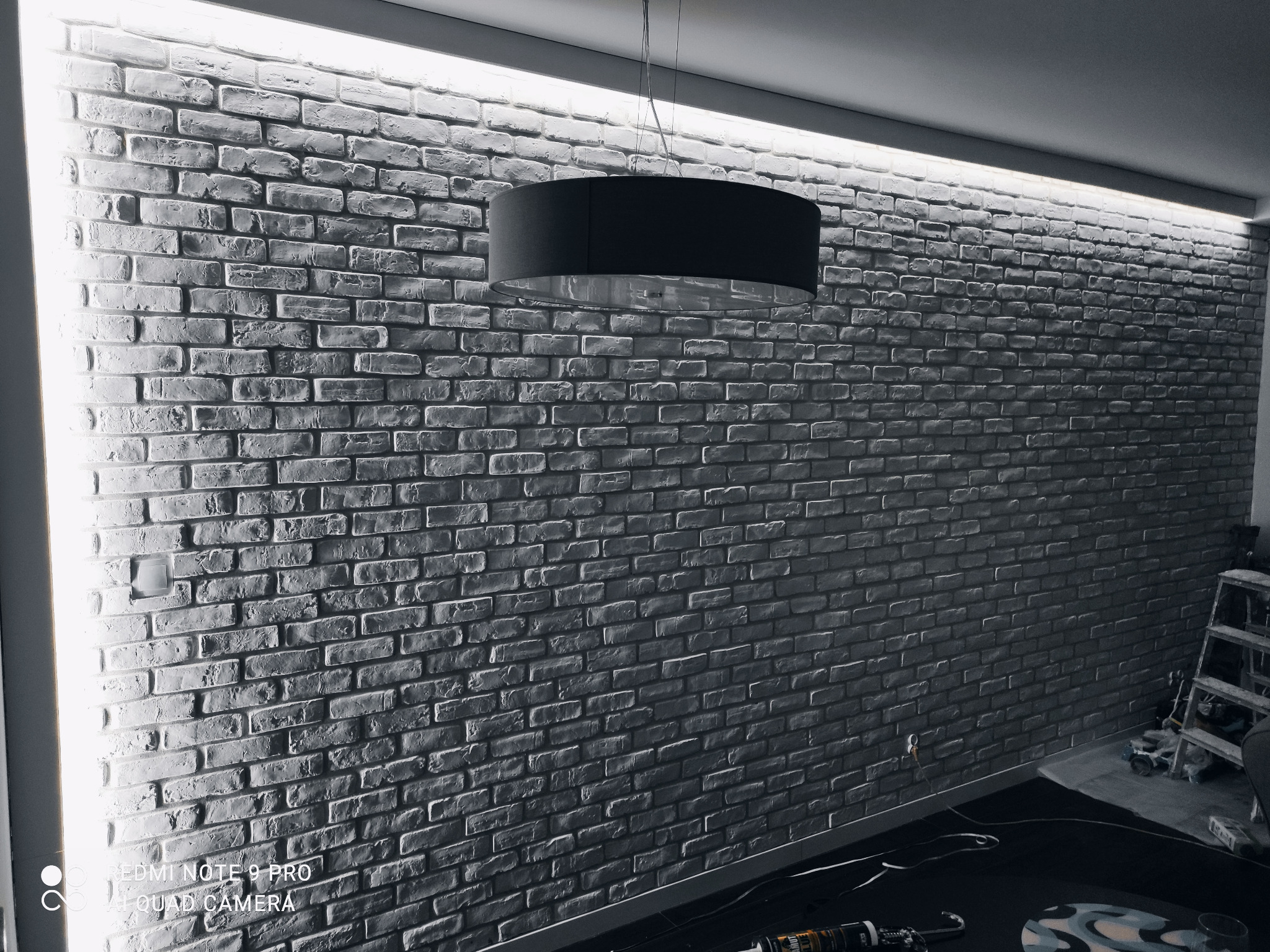 Ściana z szarej cegły z oświetleniem LED pod sufitem i wiszącą lampą z czarnym abażurem, widoczne narzędzia i drabina w tle.