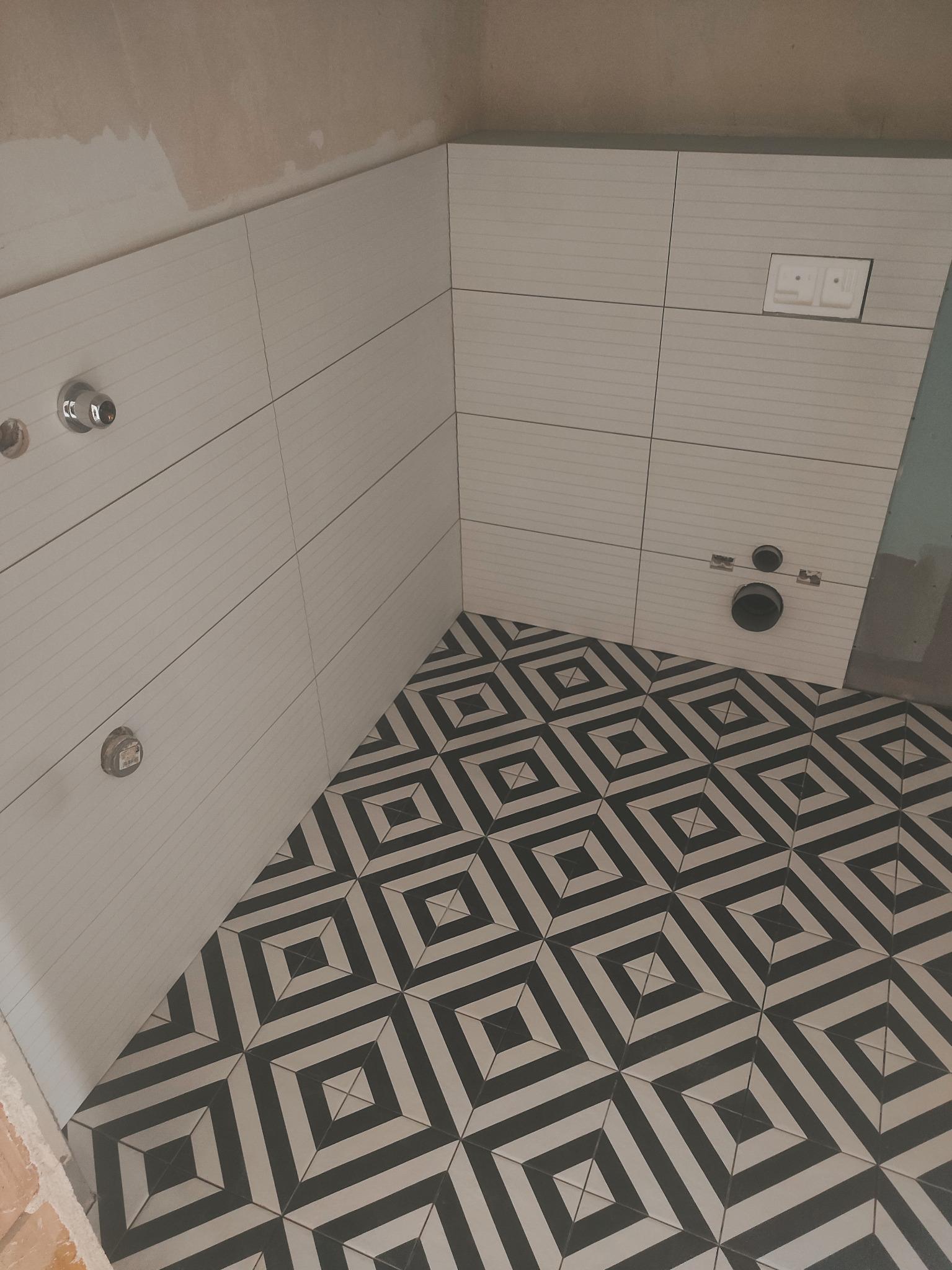 Łazienka w trakcie remontu: białe, poziome płytki ścienne z delikatnym wzorem, czarno-biała podłoga w geometryczne wzory, widoczne instalacje wodne i przycisk spłuczki.