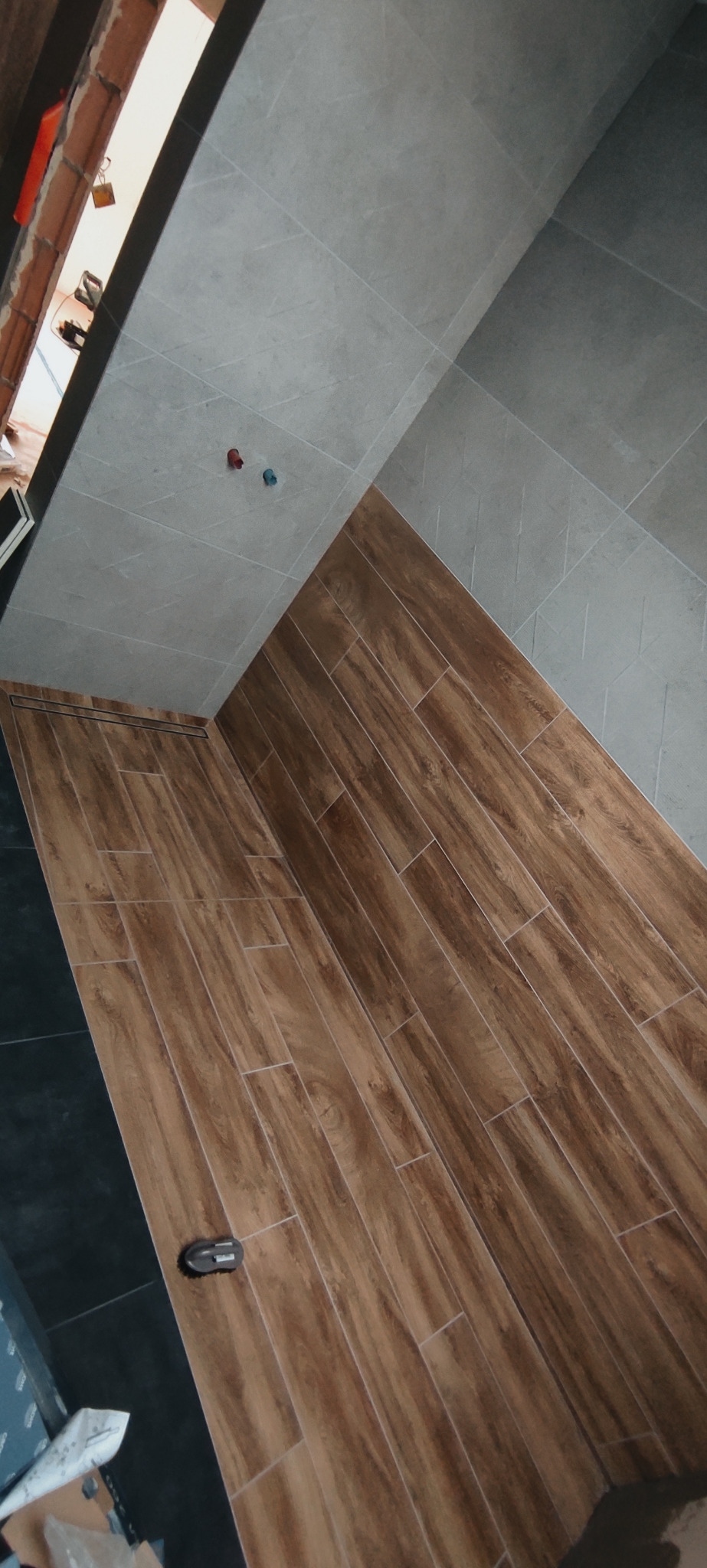 Wnętrze łazienki w trakcie prac wykończeniowych: ułożona podłoga z płytek imitujących drewno, szare płytki na ścianach, widoczne wystające rury instalacji wodnej oraz narzędzie do cięcia płytek.