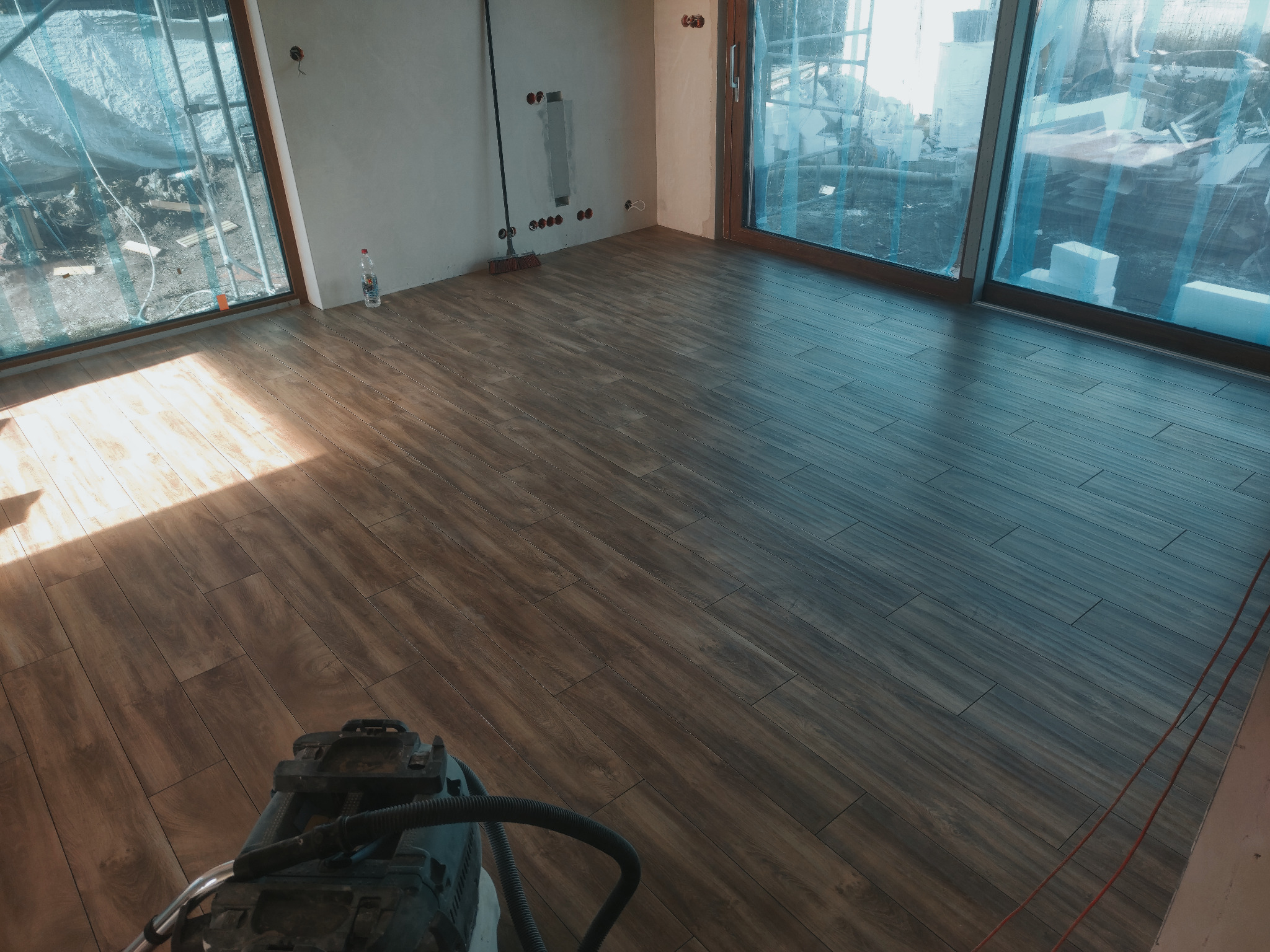 Układanie płytek imitujących drewno w jasnym pomieszczeniu z dużymi oknami, widoczna maszyna czyszcząca na podłodze i materiały budowlane za oknem.