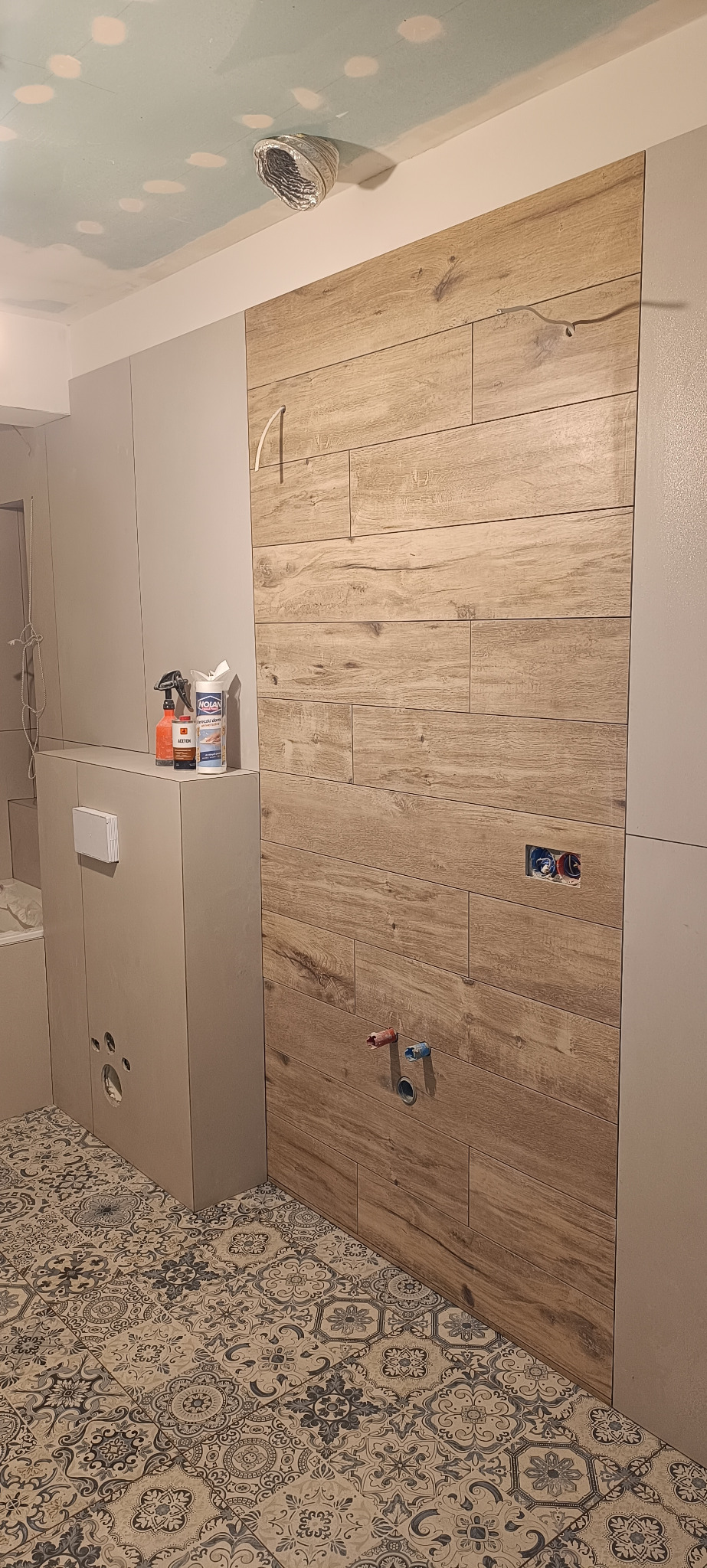 Łazienka w trakcie prac: ułożona podłoga w marokański wzór, ściana wyłożona płytkami imitującymi drewno, widoczne instalacje wodne, zabudowa stelaża WC z akcesoriami do czyszczenia na górze, surowy...