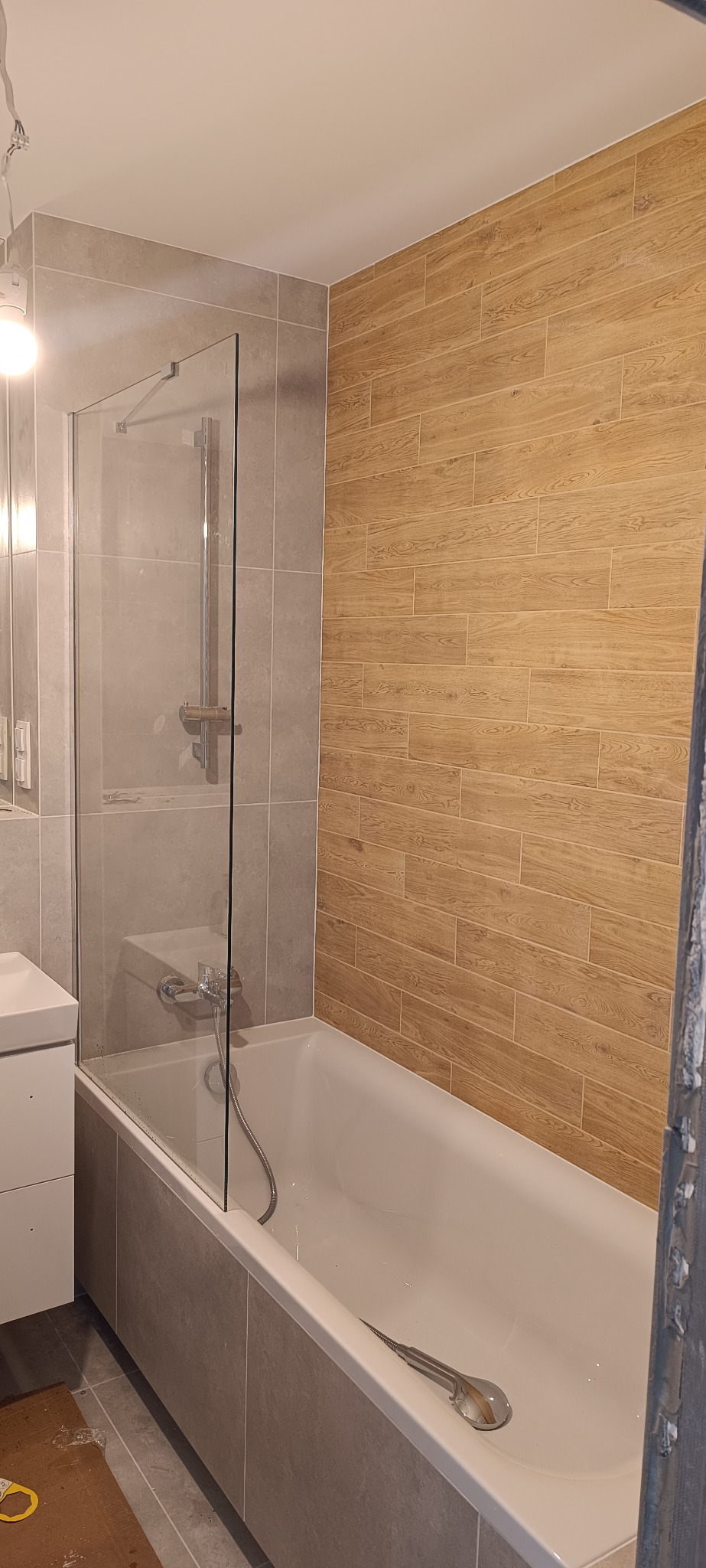 Wykończona łazienka z wanną obłożoną płytkami imitującymi kamień, szklanym parawanem nawannowym oraz ścianą wyłożoną płytkami imitującymi drewno; w wannie leży słuchawka prysznicowa.