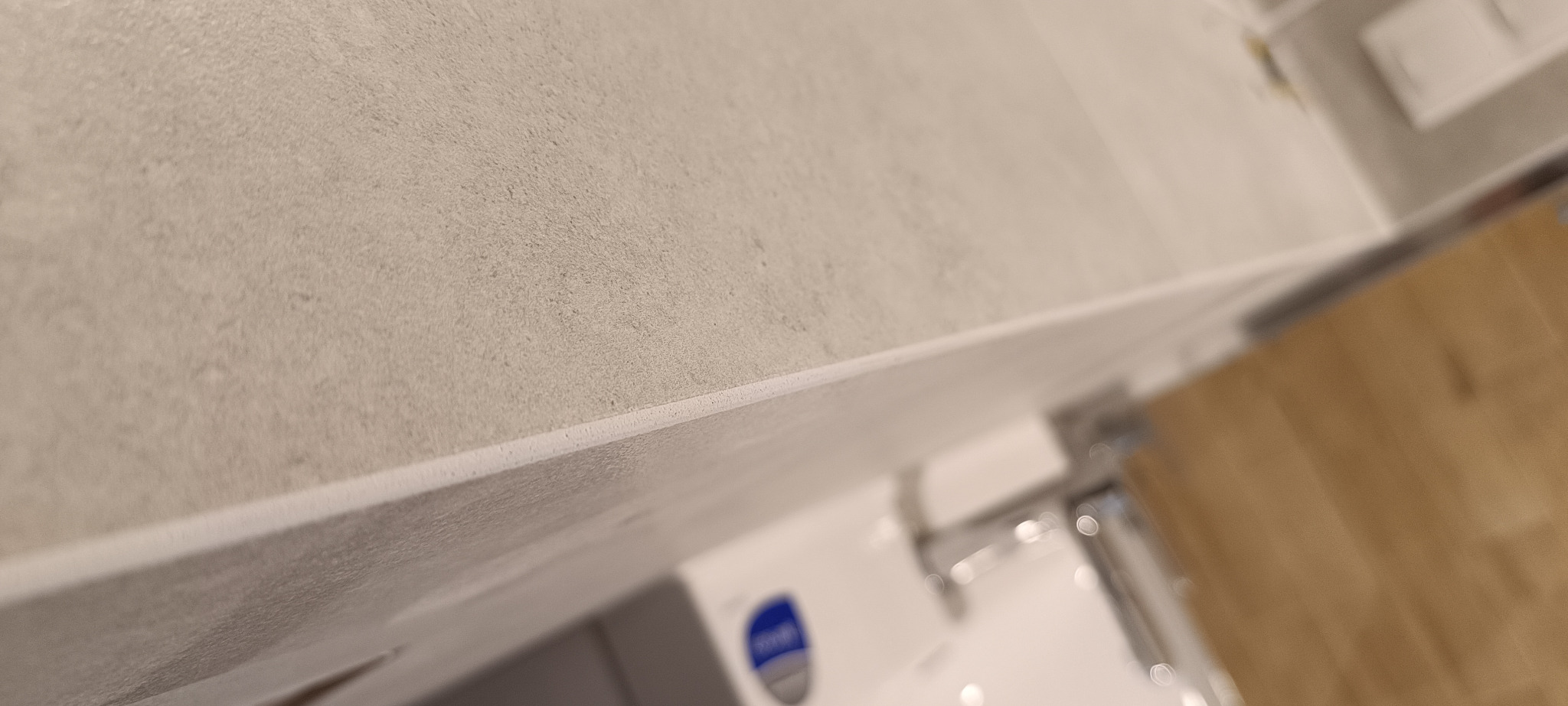 Szczegółowe ujęcie fugi między szarymi płytkami na ścianie w łazience, widoczny fragment umywalki z baterią i szafki pod umywalką.