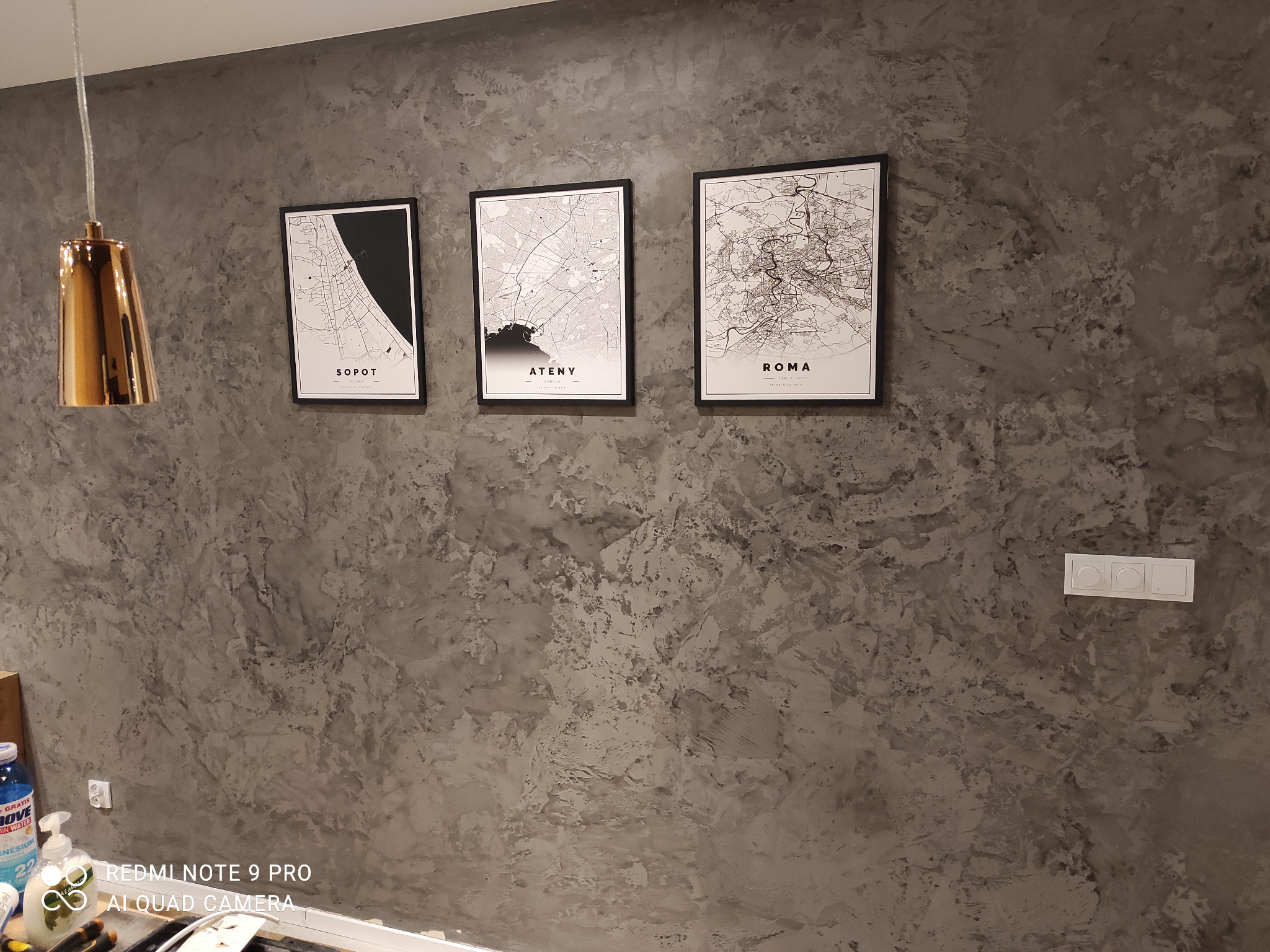 Ściana wykończona dekoracyjnym tynkiem w odcieniach szarości, z trzema czarnymi ramkami z mapami Sopotu, Aten i Rzymu oraz złotą lampą wiszącą po lewej stronie