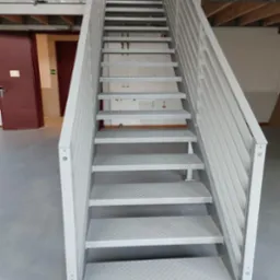 Metalowe schody przemysłowe, pomalowane na jasny kolor, z ryflowanymi stopniami, prowadzące na antresolę w hali.