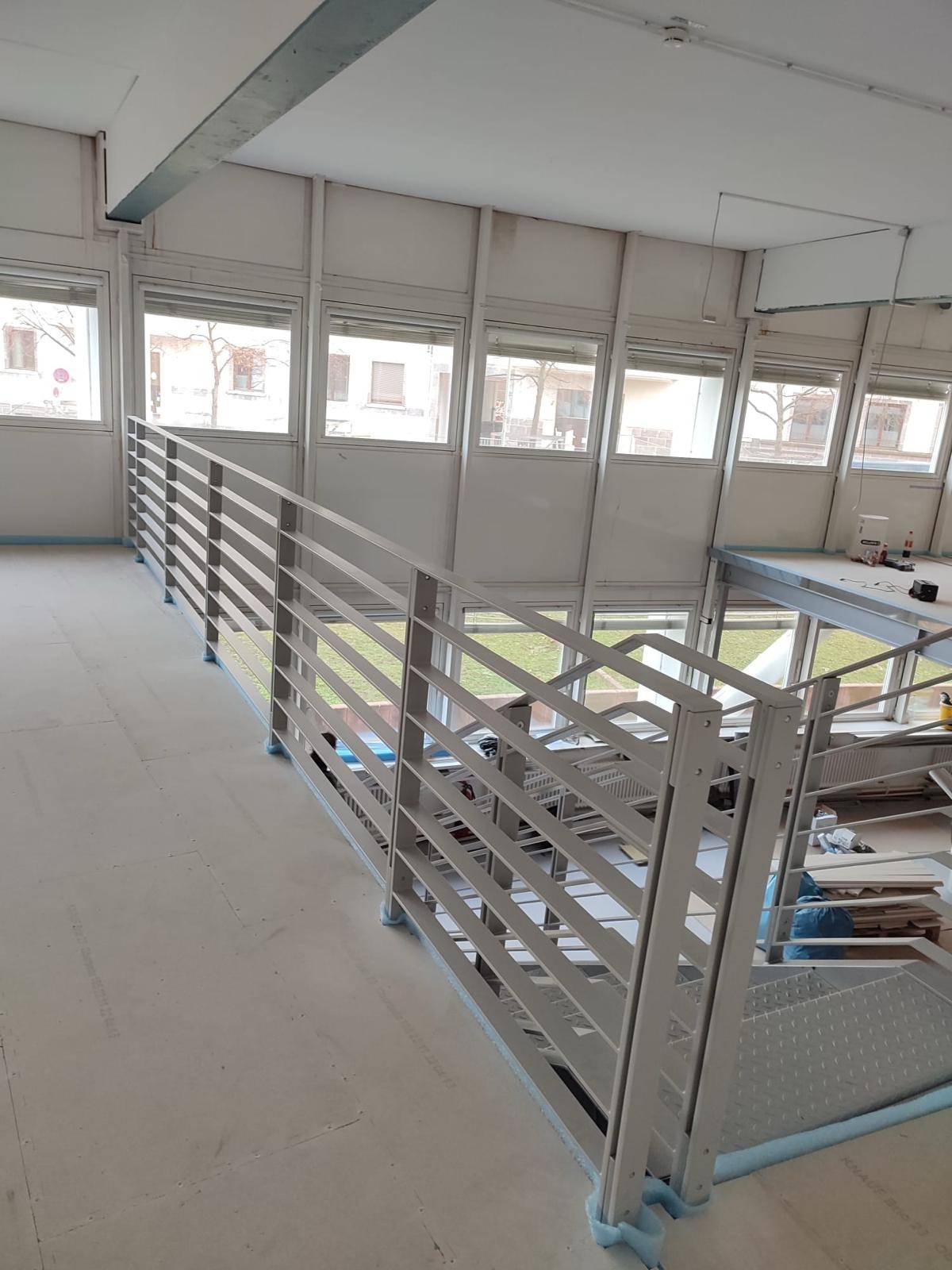 Metalowa balustrada schodowa z poziomymi szczeblami w jasnym wnętrzu budynku biurowego, widoczne okna i elementy konstrukcyjne.