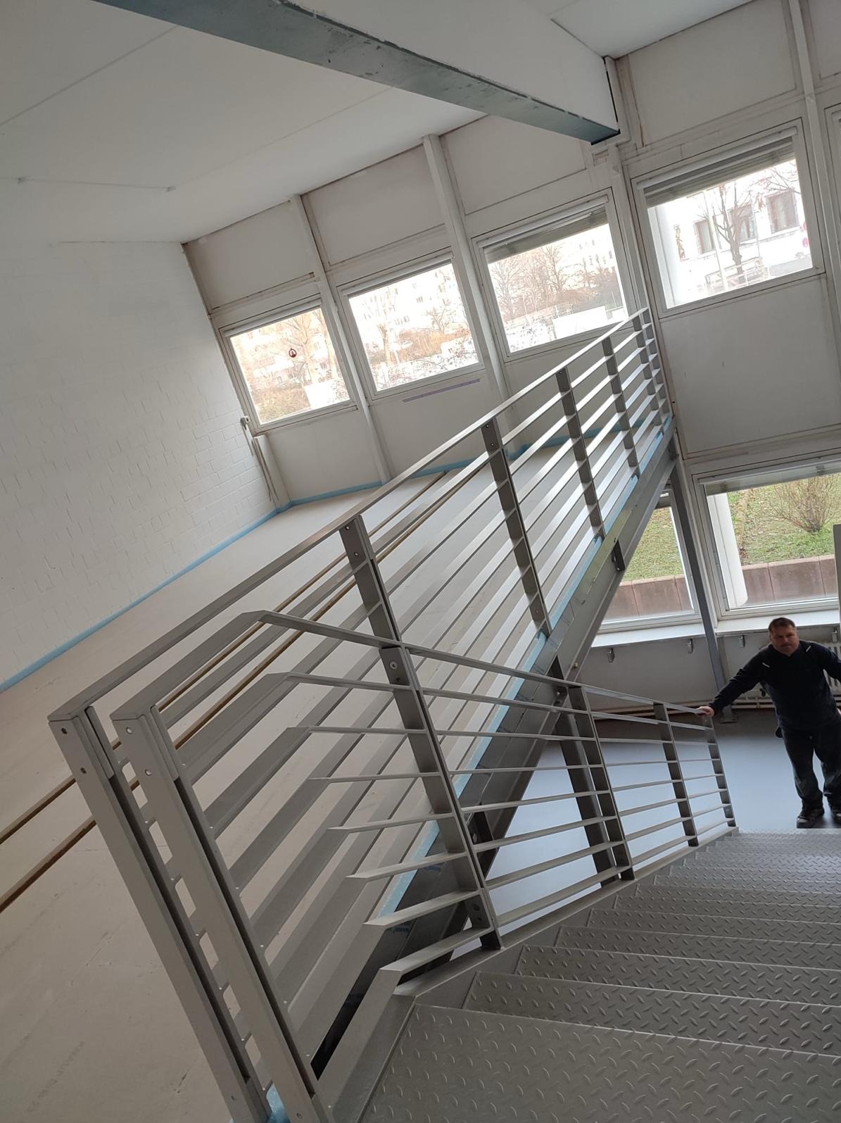 Metalowe schody z barierkami w jasnym wnętrzu budynku, widok z góry na stopnie, pracownik opiera się o barierkę.