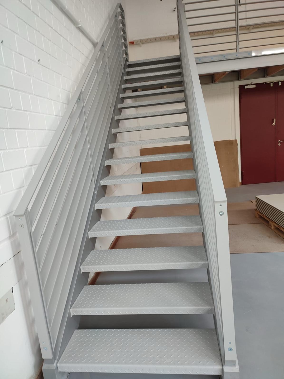Metalowe schody przemysłowe z ażurowymi stopniami i barierkami, prowadzące na antresolę w hali magazynowej. Widoczne białe ściany z cegły oraz czerwone drzwi w tle.