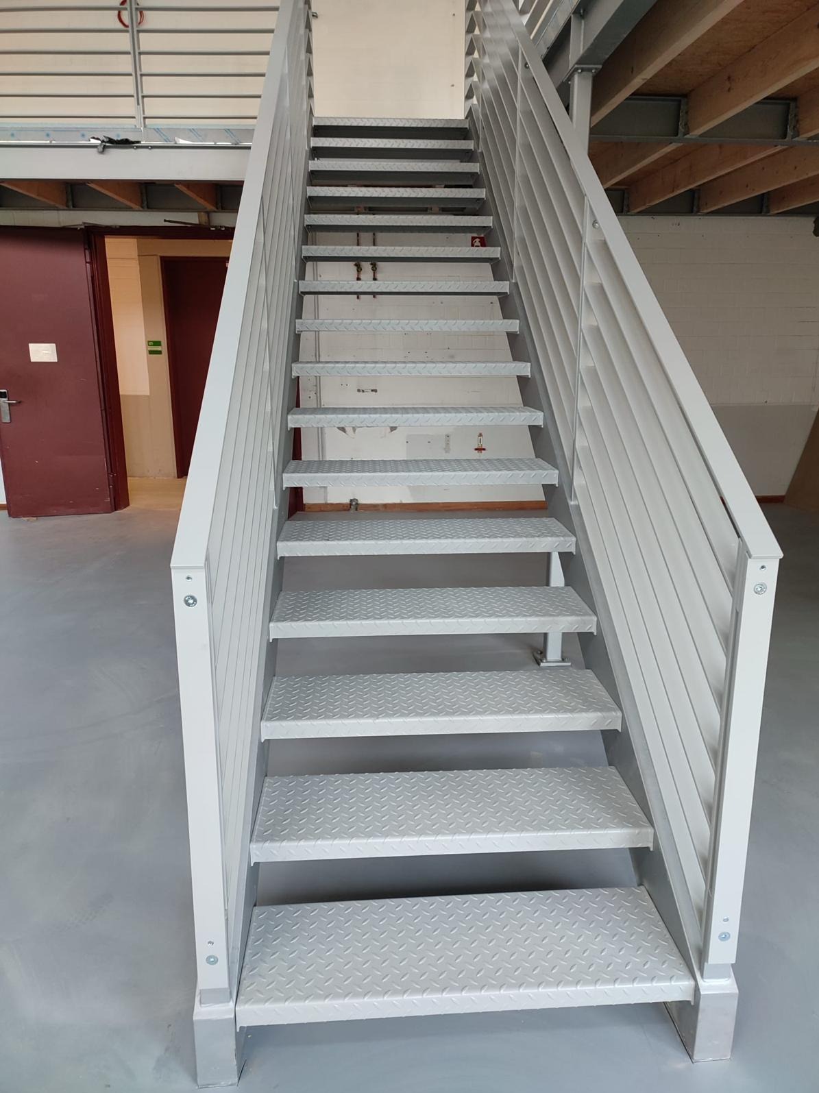 Metalowe schody przemysłowe, pomalowane na jasny kolor, z ryflowanymi stopniami, prowadzące na antresolę w hali.